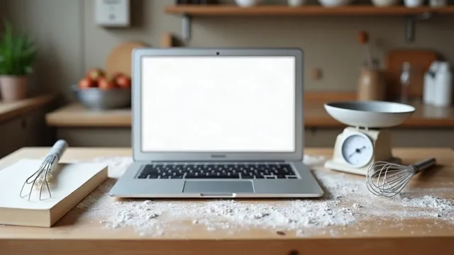An open laptop with a blank white screen on a floury wooden counter beside a kitchen scale, a whisk and a notebook.