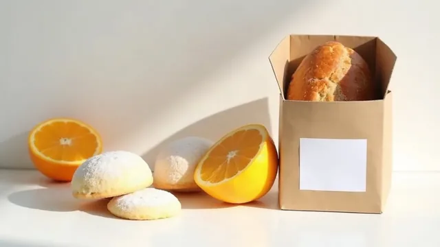 A kraft box holding a bread loaf with a blank white label, beside powdered cookies and two halved oranges on a white table.